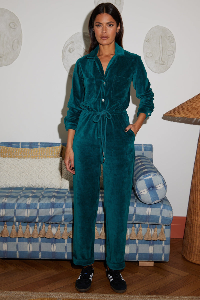 Female model faces forwards with one hand in her pocket. She wears a Dancing Leopard pine green velour jumpsuit.