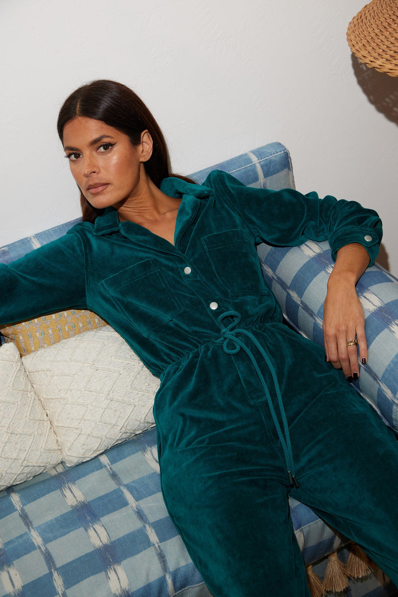 Female model sitting down on a couch. She wears a Dancing Leopard pine green velour jumpsuit.