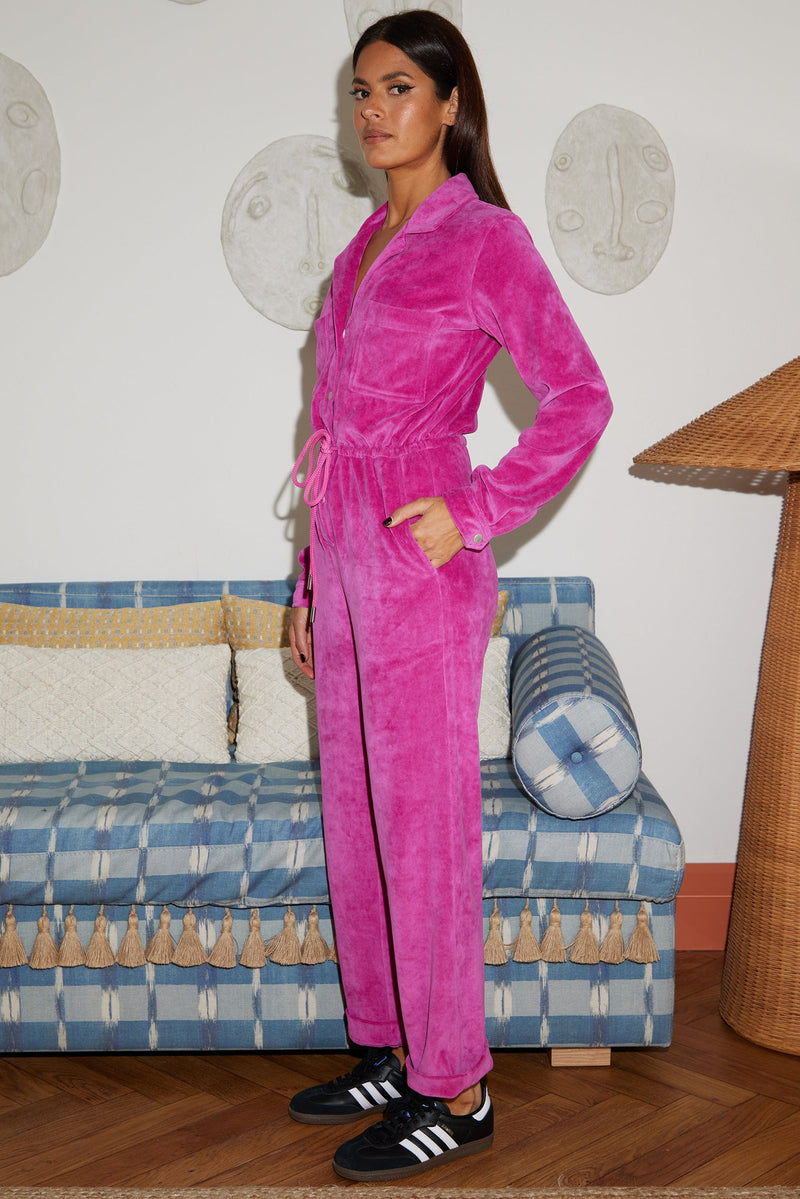 Female model faces forwards with one hand in her pocket. She wears a Dancing Leopard bright pink velour jumpsuit.