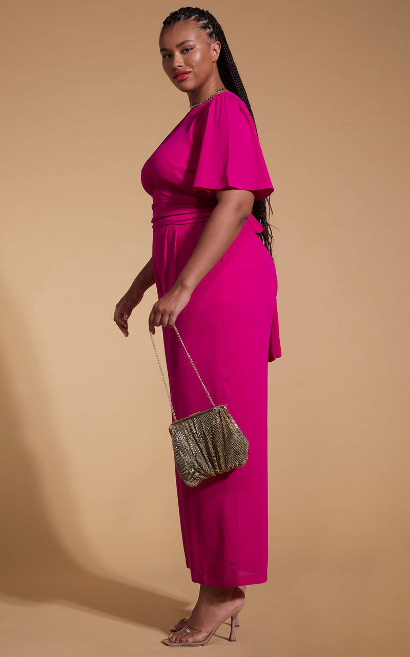 Model faces to the side wearing a pink dancing leopard jumpsuit with a gold clutch bag.