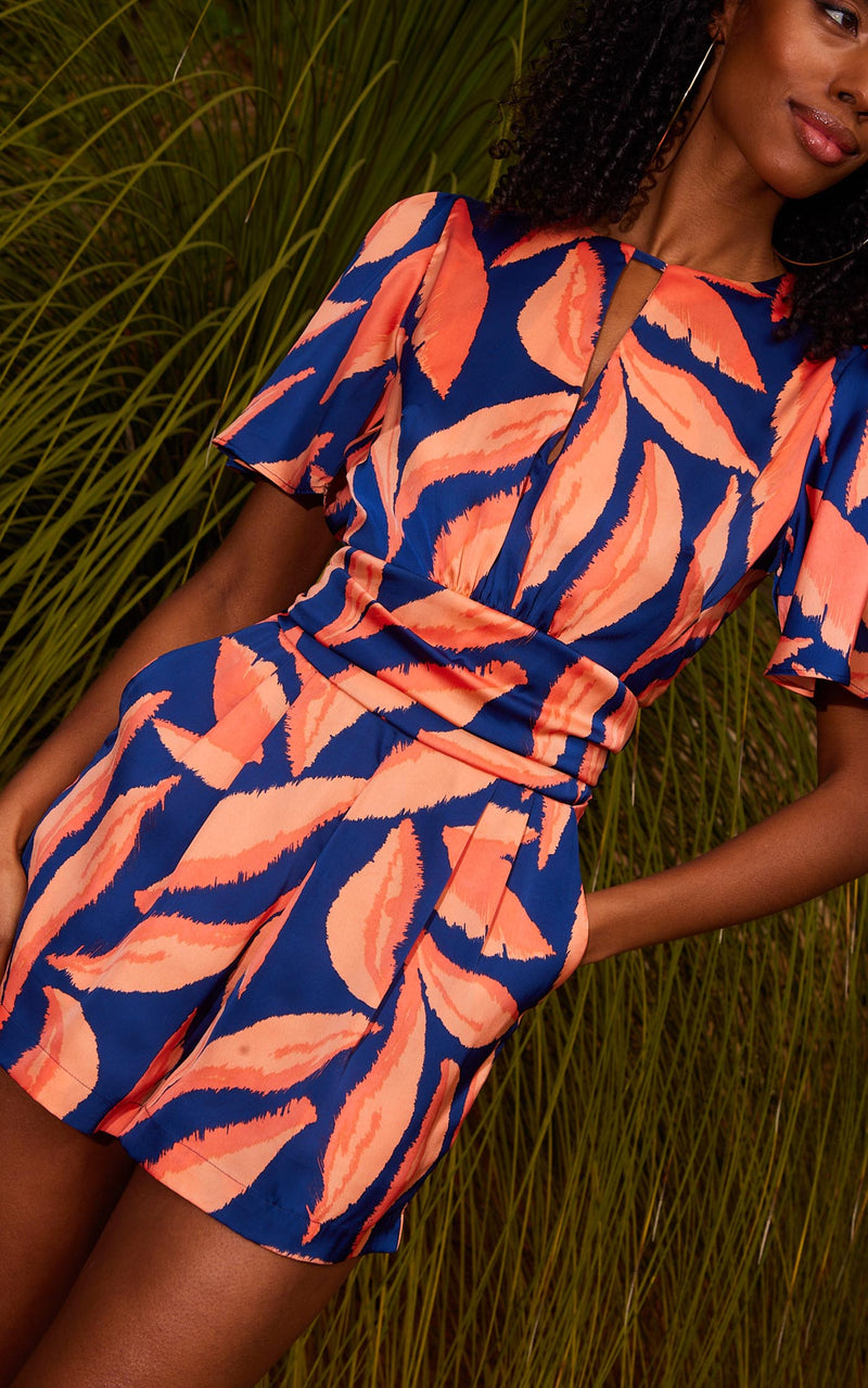 Female model faces forwards with one hand in her pocket. She wears a Dancing Leopard orange and blue abstract leaf playsuit.