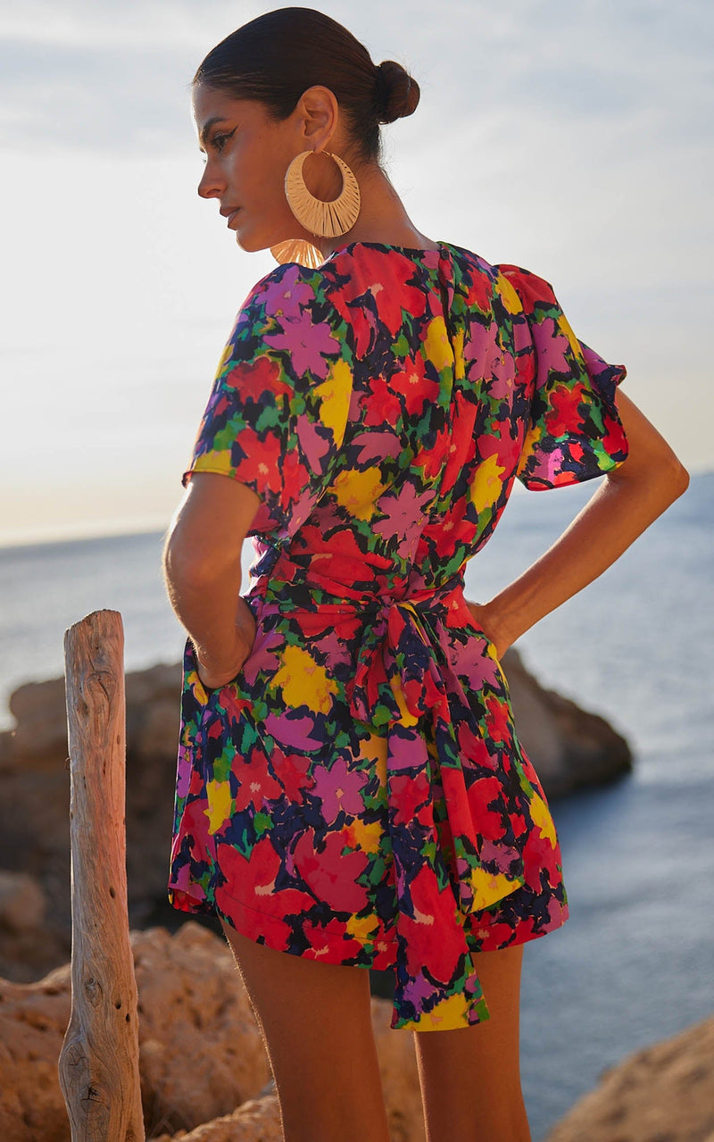 A female model faces backwards with her hands in her pockets. She wears a Dancing Leopard colourful, floral printed playsuit.