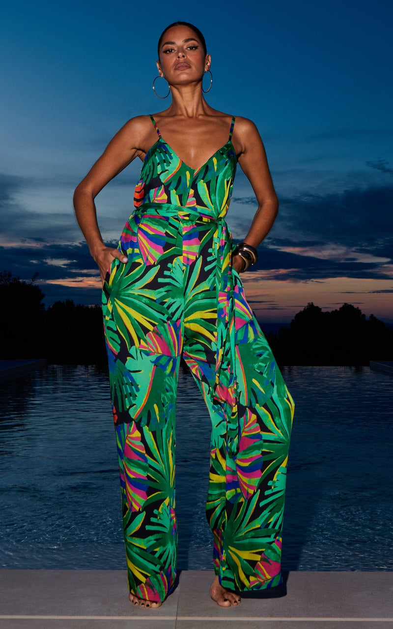 A female model faces forward in front of a pool. She wears a Dancing Leopard green, tropical print jumpsuit.
