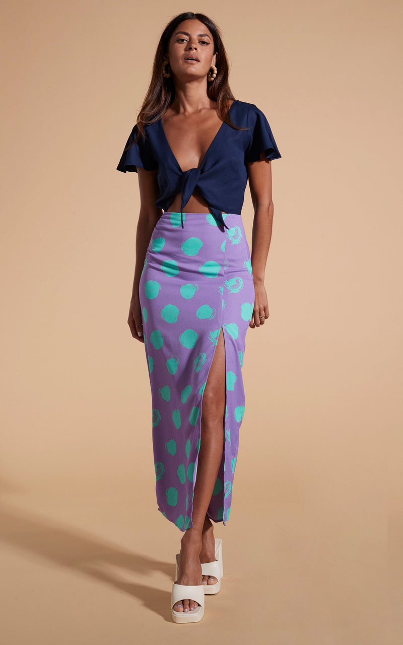 Model stands facing forwards wearing polka dot and navy maxi dress with tie-front and heels