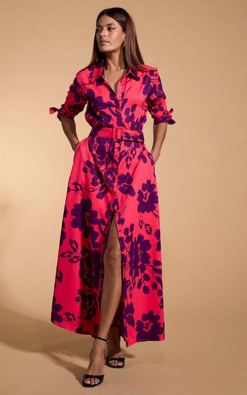 A female model faces forwards with her hands in her pockets. She wears a Dancing Leopard maxi shirt dress. The dress is red with a plum coloured flower print.