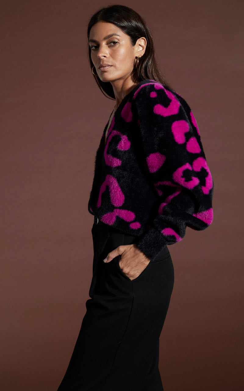 Dancing leopard model standing sideways with hands In pockets wearing bambino cardigan in cerise on black leopard with black trousers