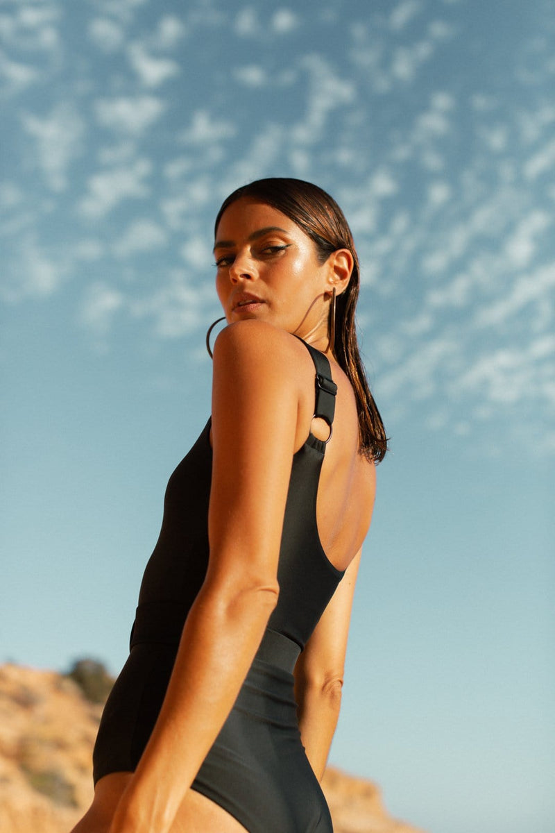 Dancing Leopard model faces side-on standing on beach wearing HALO Kiara Belted Swimsuit in Black