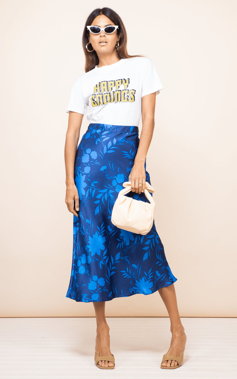 Brunette model faces forward holding bag wearing Renzo Skirt in blue floral print with sunglasses
