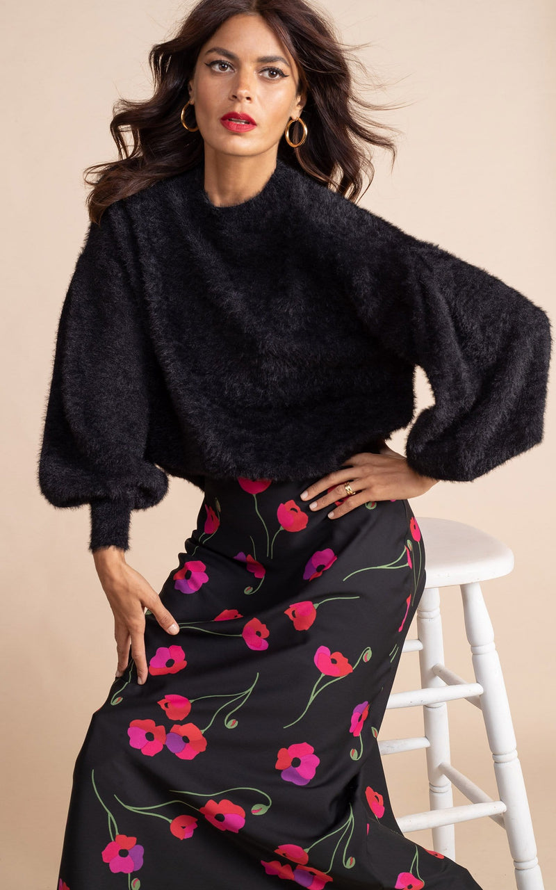 Dancing Leopard model wearing black furry jumper and black and red / pink poppies floral design slip skirt sitting on a white stool