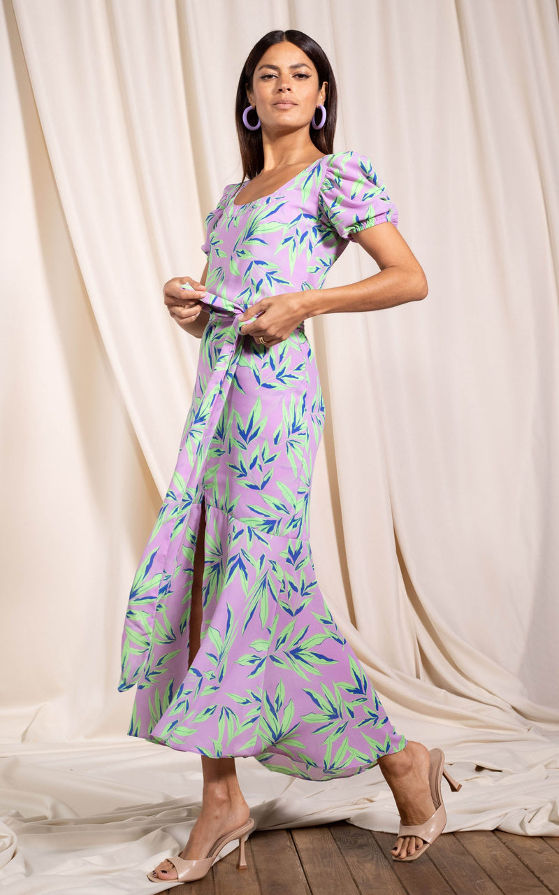 Dancing Leopard model standing sideways holding onto tie of dress wearing Manuela frill hem dress in tropic mint on pink
