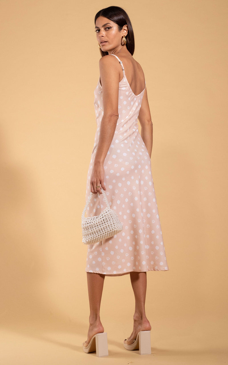 Dancing Leopard model standing with her back to the camera looking over shoulder holding woven white bag wearing Sienna midaxi dress in beige polka dot