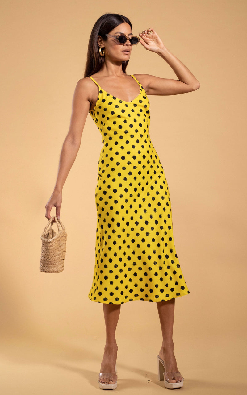 Dancing Leopard model standing wearing Sienna midaxi dress in yellow polka dot holding into straw bag and sunglasses on face