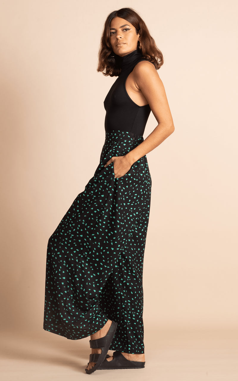 Dancing Leopard model standing sideways with hands in trouser pockets wearing wide leg black and green dotty wide leg trousers with black bodysuit and black sandals