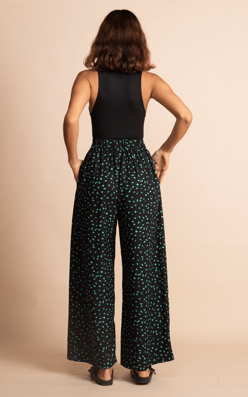 Dancing Leopard model standing with back to the camera with hands in trouser pockets wearing wide leg black and green dotty wide leg trousers with black bodysuit and black sandals