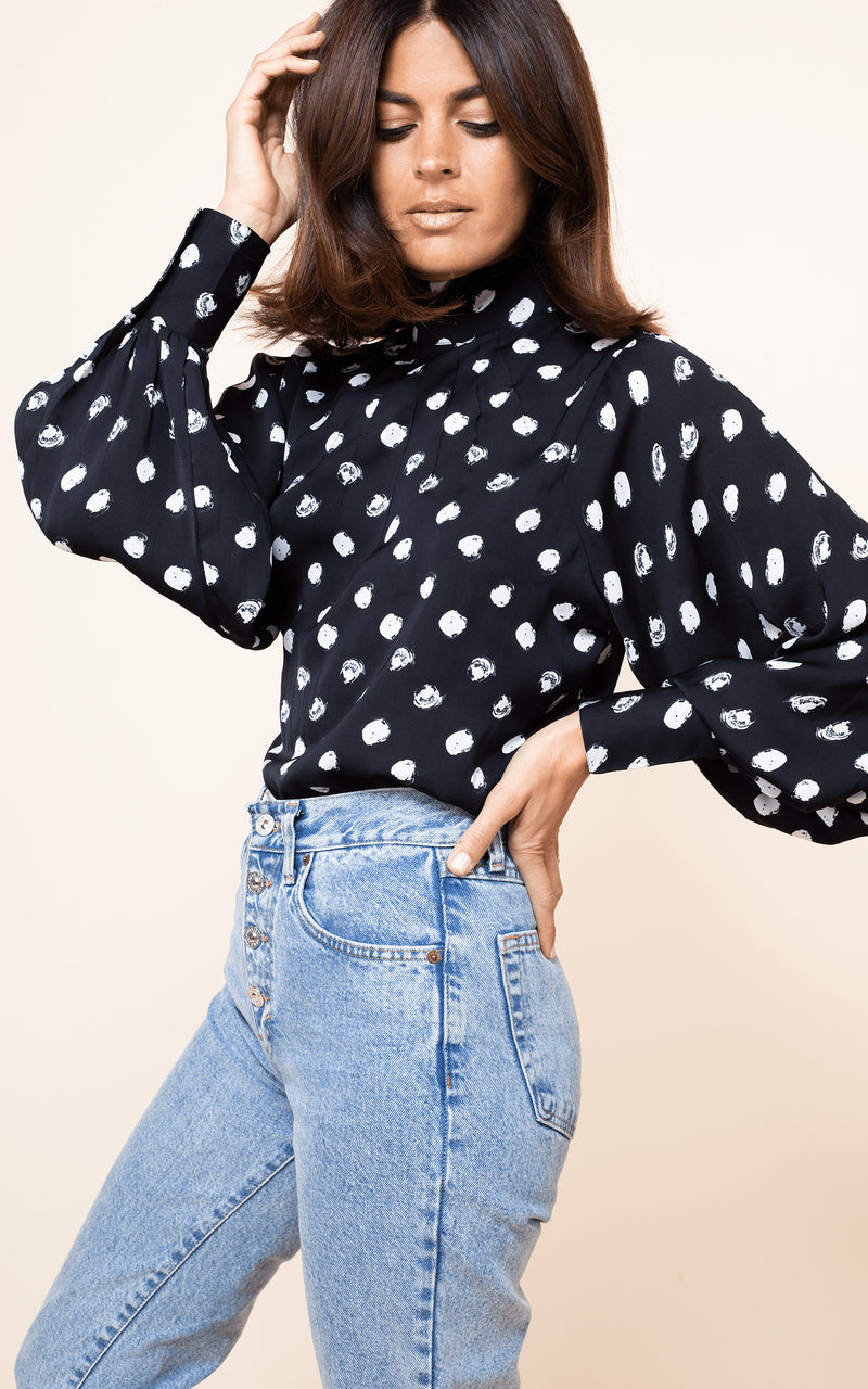Dancing Leopard model stands side-on with hand in hair wearing Margo Blouse in Painted Dot with jeans
