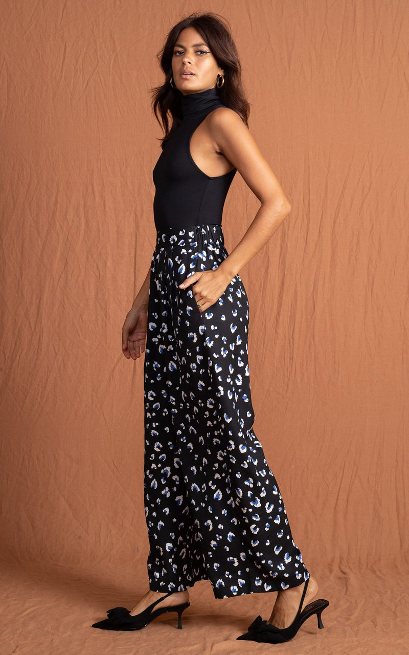 Dancing Leopard model standing sideways to the camera wearing a black top and wide leg black, white and navy blue leopard print trousers with hands in her pockets and black pumps and hand in  pocket