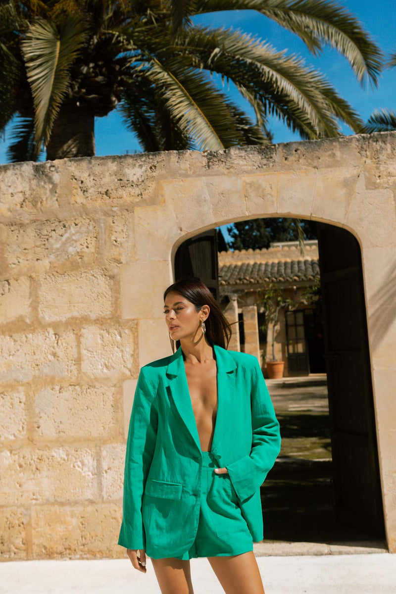 Dancing Leopard model wearing Lola Linen Shorts In Vivid Green with matching blazer in front of a wall and palm tree