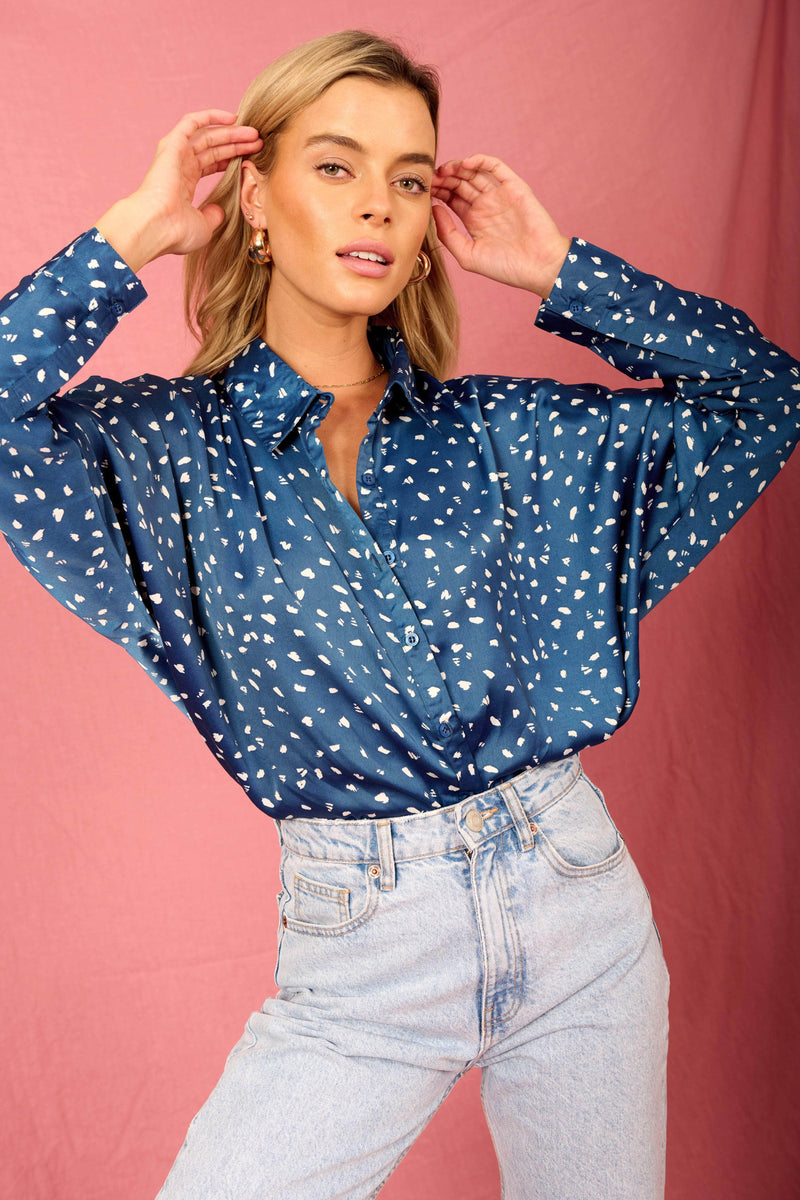 Model standing with hand against her face wearing blue and white dotty satin shirt and blue jeans