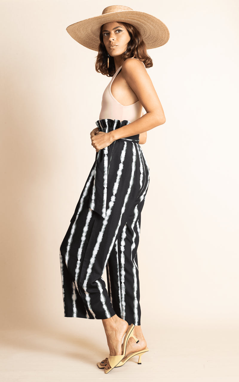 Dancing Leopard standing sideways wearing black and white stripe winnie wide leg trousers with nude bodysuit and straw hat and sandals