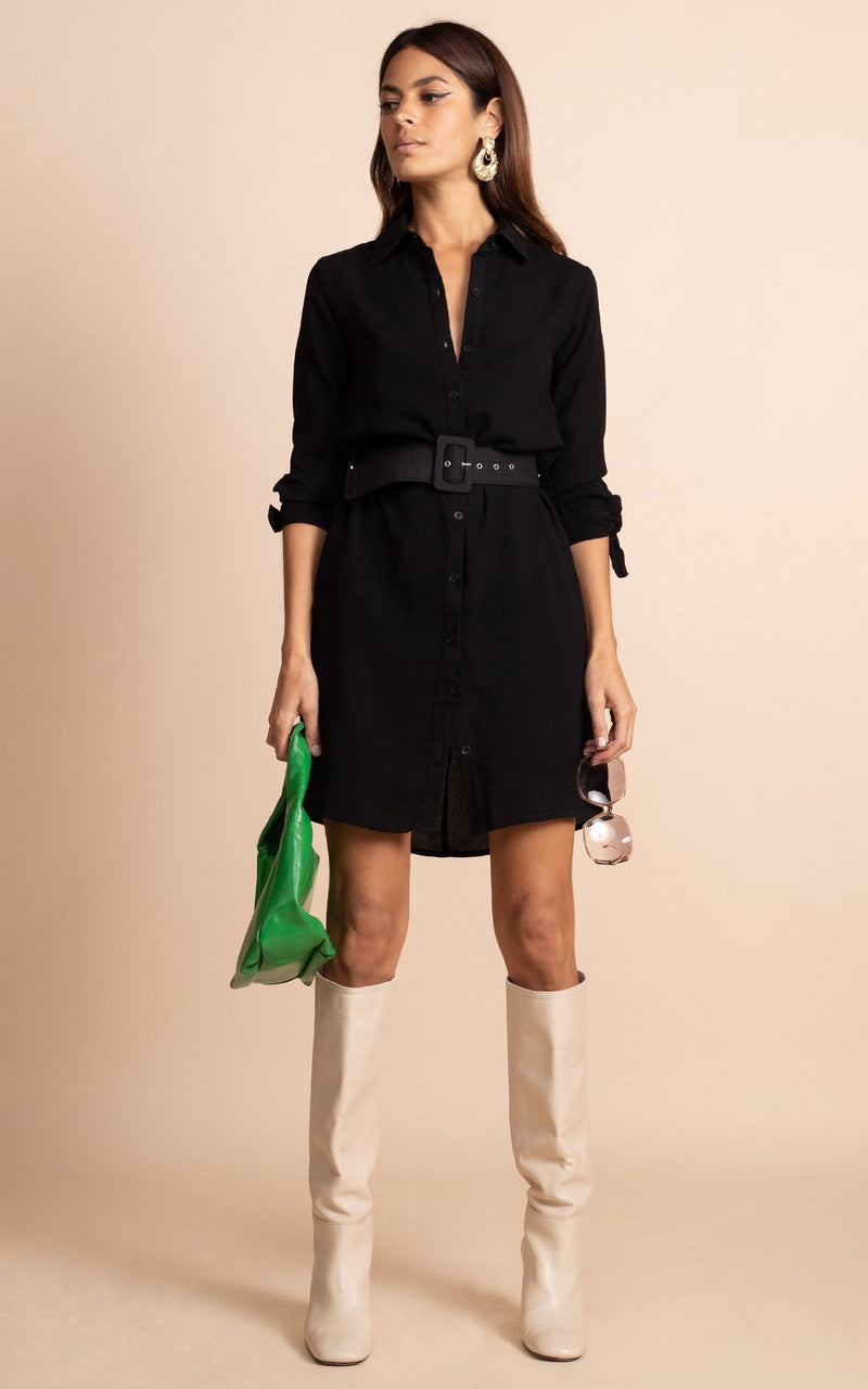 Dancing Leopard model standing face forward wearing Jonah mini skirt dress in black and holding green bag and sunglasses, with cream boots